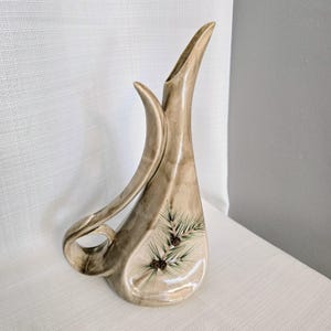 May include: A tall, beige ceramic vase with a glossy finish and an asymmetrical, elongated silhouette. The vase features a curved, sweeping handle and a painted design of green pine needles and small brown pinecones on the lower front section.
