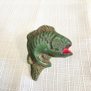 May include: A cast iron fish figurine featuring a textured green finish with visible brown rust-like accents and a bright red painted mouth. The fish is depicted in a swimming position with detailed scales and fins, showing a weathered appearance.