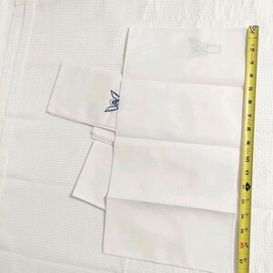 Może przedstawiać: Białe, złożone chusteczki z haftowanymi niebiesko-białymi wzorami. Ż&oacute;łta taśma miernicza jest umieszczona obok chusteczek, pokazując długość około 43 cm.