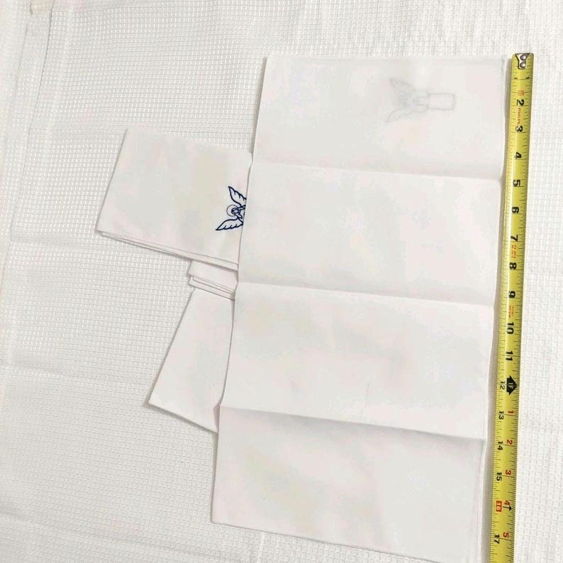 Może przedstawiać: Białe, złożone chusteczki z haftowanymi niebiesko-białymi wzorami. Ż&oacute;łta taśma miernicza jest umieszczona obok chusteczek, pokazując długość około 43 cm.