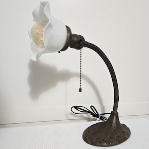 May include: A vintage-style desk lamp with a white, flower-shaped shade. The lamp features a flexible, dark brown gooseneck arm and a decorative, bronze-colored base. A pull chain hangs from the lamp's head. The lamp is designed for a warm, inviting light.