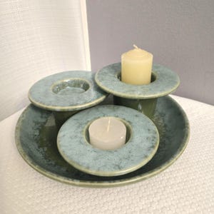May include: A ceramic candle holder set in shades of green and blue. The set includes a round base, three candle holders, and two lit candles. The candle holders have a circular design with a central opening for the candles.