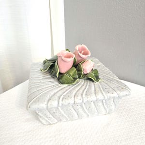May include: A square, speckled, ceramic trinket box with a lid. The lid is decorated with a cluster of three pink ceramic roses and green leaves. The box is light gray with a textured, ridged design.
