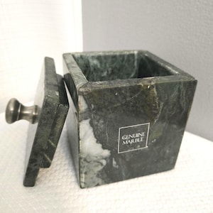 May include: A square, green marble box with a lid and a silver knob. The lid is slightly ajar, revealing the interior. The box has a "GENUINE MARBLE" sticker on the side. The marble has a natural, veined pattern.