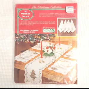 May include: A Christmas-themed napkin kit set of four, featuring a table setting with a white runner and placemats. The kit includes embroidered designs of Christmas trees and greenery. Each napkin is 14 inches square.
