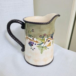 May include: A ceramic pitcher featuring a cream-colored base with a soft checkered pattern, topped with a sage green rim. The body is decorated with painted grape clusters, green leaves, and a small yellow dragonfly. It has a dark brown rope-textured handle.