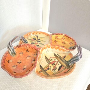 Puede incluir: Una fuente decorativa de cerámica con cuatro secciones y un asa. La fuente presenta diseños pintados a mano de aceitunas, flores y edificios en tonos naranja, amarillo y verde. El borde tiene un borde festoneado.