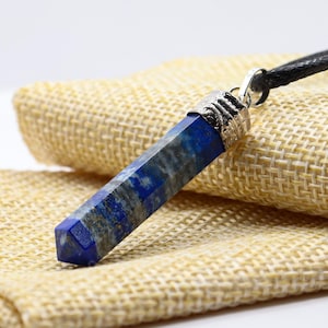 Op de afbeelding: Een blauwe en grijze lapis lazuli hanger ketting. De edelsteen heeft een rechthoekige prisma vorm met een zilverkleurige metalen dop. De ketting heeft een zwart koord. De hanger wordt gepresenteerd op een beige stof.