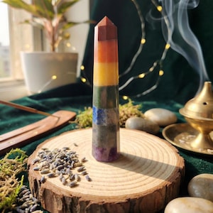 Pode incluir: Uma torre de cristal multicolorida com um topo pontiagudo, mostrando camadas de marrom, laranja, amarelo, verde, azul e roxo. O cristal está em uma fatia de madeira com lavanda seca e musgo. A fumaça do incenso sobe ao fundo.