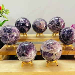 May include: Seven polished amethyst spheres with deep purple and white crystalline patterns, each resting on a small, ornate gold-toned metal tripod stand. The spheres are arranged on a tiered wooden display shelf with a smooth, light-colored finish.
