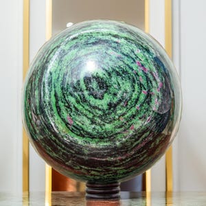 May include: A polished spherical stone ornament featuring a vibrant pattern of swirling green and black mineral deposits with scattered inclusions of deep pink and magenta. The sphere rests on a small, dark, circular tiered base stand.