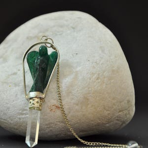 May include: A pendulum with a silver-colored metal frame and chain. The pendulum features a carved green angel figure and a clear crystal point. The chain ends with a small clear crystal ball. The background is a white stone.