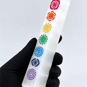 Peut inclure: Une tour en cristal de sélénite blanc translucide à section hexagonale, présentant sept symboles de chakra alignés verticalement et imprimés en rouge, orange, jaune, vert, bleu clair, bleu foncé et violet sur un côté.