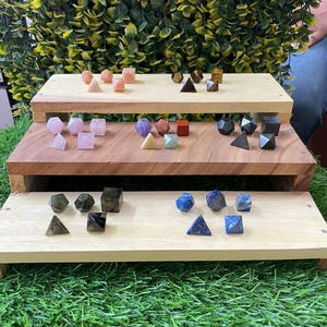 Könnte beinhalten: Eine Sammlung kleiner, polierter geometrischer Steinformen, darunter Pyramiden, Würfel und Polyeder, angeordnet auf einem dreistufigen Holzständer. Die Steine weisen verschiedene Farben wie Rosa, Violett, Blau, Grün, Braun und Schwarz auf.