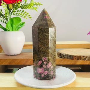 Op de afbeelding: Een gepolijste zeshoekige kristaltoren met een donker olijfgroene en bruine basis met ingebedde levendige magenta en roze minerale insluitingen. De steen heeft een glad, reflecterend oppervlak en een puntige bovenkant op een wit bord.