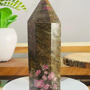 May include: A polished hexagonal crystal tower featuring a dark olive green and brown matrix base with embedded vibrant pink ruby inclusions. The stone has a smooth, reflective surface and a pointed top, showcasing natural mineral textures throughout.
