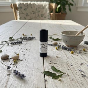 Op de afbeelding: Een zwart cilindrisch product met een wit etiket, getiteld "Tallow Cleo", staat op een wit houten oppervlak. Lavendel takjes, kamille bloemen en kleine stenen zijn verspreid rond het product, wat wijst op een natuurlijk huidverzorgingsthema.