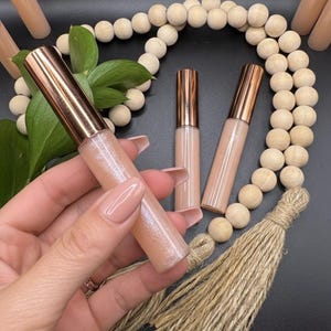 May include: Lip gloss tubes with rose gold lids, filled with a shimmery, pale pink gloss. The tubes are arranged with wooden beads, greenery, and a jute tassel against a dark backdrop.