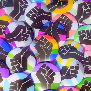 May include: A collection of colorful round buttons with a raised fist design. The buttons feature various pride flag designs, including the transgender, bisexual, and pansexual flags.