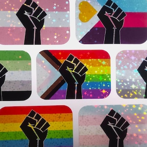 May include: A selection of stickers displaying a black fist graphic over LGBTQ+ pride flag designs. The stickers have a holographic, glittery finish, with a spectrum of colours and patterns.