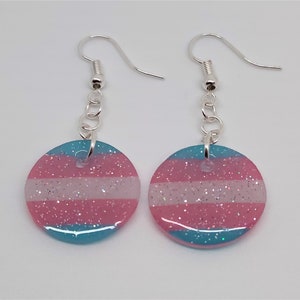 May include: A pair of silver dangle earrings with a round, blue, pink, and white glitter charm. The charm features the transgender pride flag colors.