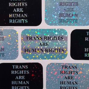 Könnte beinhalten: Ein Bogen Aufkleber mit dem Text "TRANS RIGHTS ARE HUMAN RIGHTS" in einer holografischen, glitzernden Schrift. Die Aufkleber sind rechteckig mit abgerundeten Ecken, und der Hintergrund hat einen blauen und schwarzen Farbverlauf.