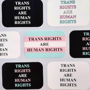 May include: A collection of stickers with the text "TRANS RIGHTS ARE HUMAN RIGHTS". The stickers are in various colors, including black, white, and light blue, with the text in a sans-serif font. Some stickers feature the transgender pride flag.