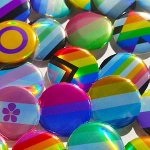 Könnte beinhalten: Eine Sammlung bunter, runder Knöpfe mit verschiedenen Designs, darunter eine Regenbogenflagge, eine Transgenderflagge, ein Friedenszeichen und eine Blume. Die Knöpfe sind aus einem glänzenden, schillernden Material gefertigt.