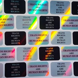 Pode incluir: Folha de autocolantes com o texto "TRANS RIGHTS ARE HUMAN RIGHTS". Os autocolantes retangulares com cantos arredondados apresentam um acabamento holográfico ou iridescente, refletindo várias cores.