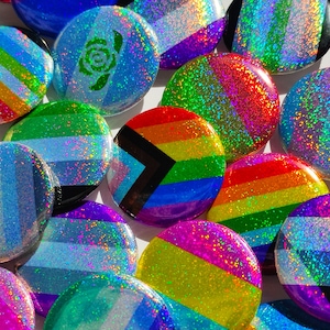 Puede incluir: Una colección de botones de solapa brillantes y coloridos que representan diversas banderas del orgullo, incluidas las banderas transgénero, bisexual, lesbiana y pansexual. Los botones son todos redondos y tienen un acabado brillante e iridiscente.