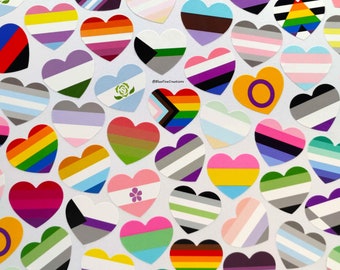 May include: A collection of heart-shaped stickers featuring various pride flags, including the rainbow, transgender, bisexual, lesbian, and pansexual flags. The stickers are arranged in a random pattern on a white background.