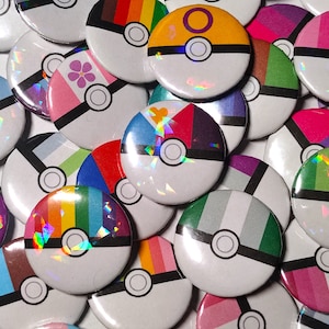 Könnte beinhalten: Eine Sammlung bunter, runder Knöpfe mit weißem Hintergrund und einem schwarzen Kreis in der Mitte. Jeder Knopf zeigt ein anderes Design einer Pride-Flagge, darunter die Regenbogen-, Bi-, Transgender-, Lesben- und Genderfluid-Flaggen.