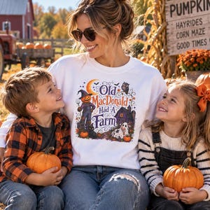 May include: A white long-sleeve sweatshirt featuring a colorful graphic print with the text "Old MacDonald Had A Farm," a pumpkin-headed figure, a barn, a cow, and an owl, worn by a person sitting on hay with two children holding small pumpkins.