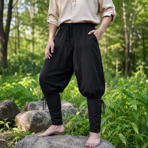 May include: Black trousers featuring a loose, baggy fit through the thighs and a drawstring waistband. The lower legs are fitted with a lace-up design that includes metal eyelets and black cord ties, creating a distinct silhouette at the ankles.
