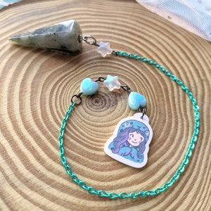 Puede incluir: Una cadena verde azulada con una punta de cristal transparente, un dije de una niña con un suéter azul con capucha y dijes de corazón y estrella blancos.