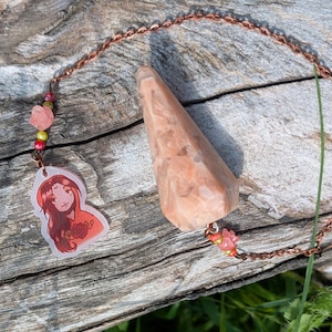 Può includere: Un pendolo per la divinazione con una catena di rame e un cristallo a forma di cono color pesca. La catena è adornata con perline rosa a forma di rosa e un ciondolo con una donna dai capelli rossi e una rosa.