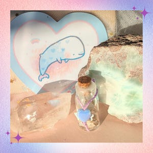 Peut inclure: Une petite bouteille en verre avec un bouchon en liège, ornée d'un cœur bleu et d'une ficelle violette. La bouteille est posée sur une surface rose, à côté d'une carte en forme de cœur avec une illustration de baleine. Des cristaux et des roches sont également présents.