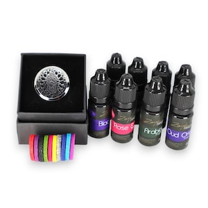 Puede incluir: Un conjunto de ocho botellas de 20 ml de mezclas de aceites esenciales para difusores. Las botellas son negras con etiquetas plateadas y presentan diferentes aromas, incluyendo Black, Rose & Oud, Arabic, Oud y Orange. El conjunto también incluye un medallón difusor plateado y una caja de almohadillas de fieltro de colores.