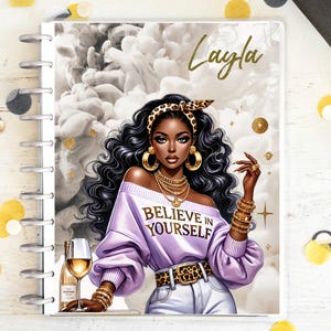 Può includere: Un'agenda a spirale con una copertina raffigurante l'illustrazione di una donna con capelli ricci scuri, gioielli d'oro e una fascia e cintura leopardata. Il testo "Believe in Yourself" è sulla sua maglia lilla. Il nome "Layla" è scritto in oro.
