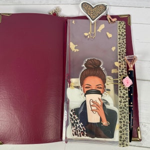 Puede incluir: Una agenda de cuero burdeos con un bolsillo transparente de plástico en la parte delantera. El bolsillo contiene una pegatina de una mujer con cabello castaño sosteniendo una taza de café. La pegatina está decorada con un estampado de leopardo. La agenda tiene un cierre de metal dorado y un anillo de metal dorado.
