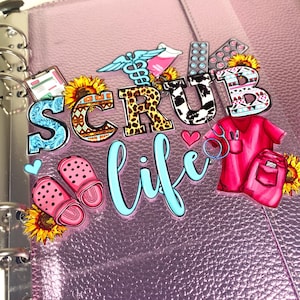 May include: A pink planner with the words "SCRUB life" in a mix of fonts and patterns. Decorative stickers include medical symbols, sunflowers, pink clogs, and a red scrub top and pants.