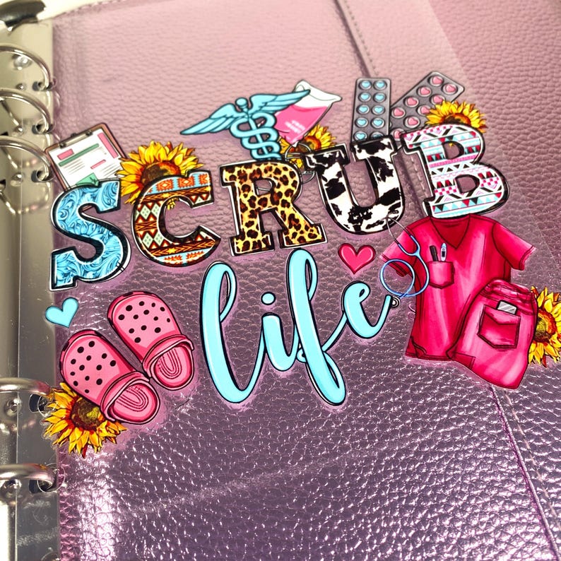 May include: A pink planner with the words "SCRUB life" in a mix of fonts and patterns. Decorative stickers include medical symbols, sunflowers, pink clogs, and a red scrub top and pants.