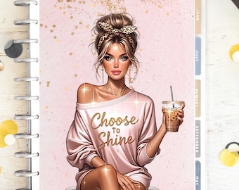May include: A planner with a pink cover featuring an illustration of a woman in a pink sweater holding an iced coffee. The text "Choose to Shine" is written on the sweater in gold glitter. The planner has monthly tabs.