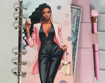 May include: A pink leather planner with a decorative insert featuring a fashion illustration of a woman in a black jumpsuit and pink blazer. A ten-dollar bill is visible, along with a pink pen.