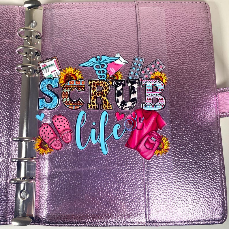May include: A purple binder with silver rings and the words "SCRUB LIFE" in colorful letters. The binder has decorative images of medical symbols, sunflowers, pink clogs, and a pink scrub top.