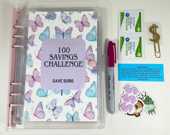 May include: A clear plastic binder with a butterfly pattern and the text "100 Savings Challenge Save 5,050" on the cover. The binder is open and there are several items next to it, including a pink highlighter, two alcohol prep pads, a gold paperclip, and a dollar sign sticker.