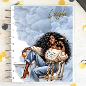 Può includere: Un'agenda rilegata a spirale con una copertina raffigurante l'illustrazione di una donna con capelli neri ricci, gioielli d'oro e un top bianco con la scritta "CREATING MY OWN PATH". Lo sfondo è un disegno di nuvole azzurre, con il nome "Layla" in oro.