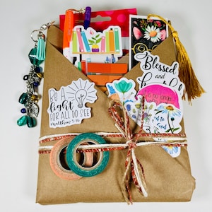 Może przedstawiać: Brązowy, papierowy pakiet prezentowy związany sznurkiem i taśmą washi. Opakowanie zawiera naklejki, długopisy i zakładki. Opakowanie jest ozdobione naklejkami z napisem „Be a light for all to see” i „Blessed and Do”. Dołączony jest brelok z zielonymi koralikami i frędzlem.