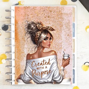 Può includere: Un'agenda con un'illustrazione di una donna bionda con gli occhi azzurri, che indossa una maglia bianca con spalle scoperte con la scritta "Created with a Purpose". Tiene in mano una tazza di caffè. Lo sfondo è rosa e oro.