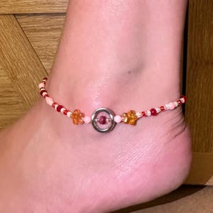 May include: Anklet featuring a beaded design with red, orange, and white beads. The anklet includes star and flower-shaped beads, and a silver-colored circular centerpiece. The anklet is worn on an ankle.